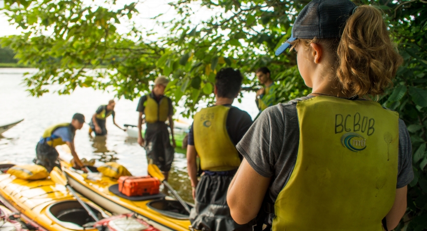 a group of young people wearing life jackets stand near kayaks beached on the shore 